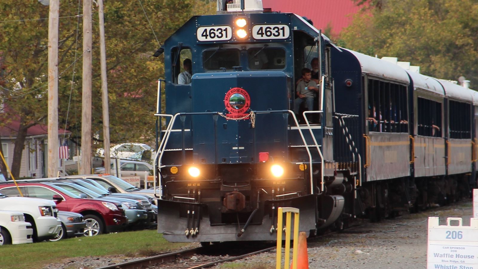 A blue Blue Ridge Scenic Railway locomotive pulling passenger cars past parked cars in Blue Ridge, Georgia
